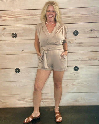 Woman wearing a beige romper standing against a wooden wall.