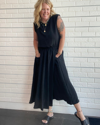 A woman modeling a sleeveless black midi dress with side pockets, standing against a white brick wall.