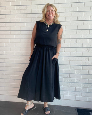 A woman wearing a black sleeveless midi skirt top set with side pockets, standing against a white brick wall.