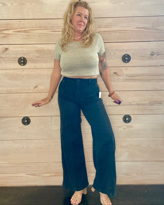 Woman wearing a light green crop top and black jeans standing against a wooden wall.