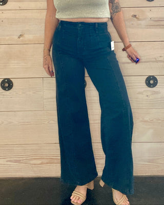 Woman wearing a light green top and black jeans standing against a wooden wall.