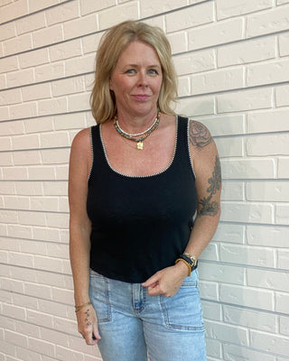 Woman wearing a black tank top and blue jeans standing against a light brick wall.