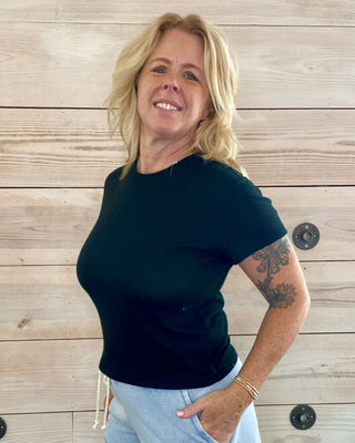 Woman wearing a black t-shirt and light blue shorts standing against a wooden wall.