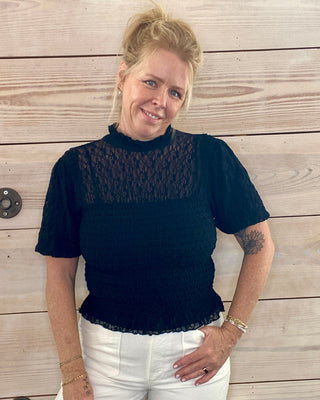 Woman wearing a black top with lace details and white pants standing against a wooden wall.