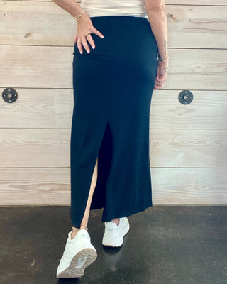 Person wearing a white sleeveless top and  black midi skirt standing against a wooden wall.
