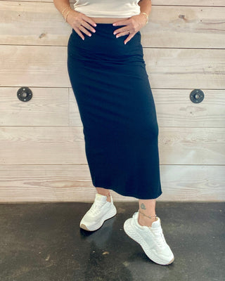 Woman wearing a white sleeveless top and black skirt against a wooden wall.
