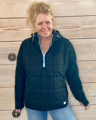 Woman wearing a black puffer jacket and blue jeans standing against a wooden wall.