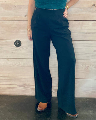 Woman wearing a green top and black knit pants standing against a wooden wall.