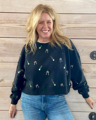 Woman wearing a black sweater with small champagne patterns and blue jeans standing against a wooden wall.