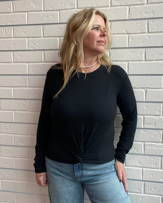 Person wearing a black long-sleeve top and blue jeans standing against a light gray brick wall.