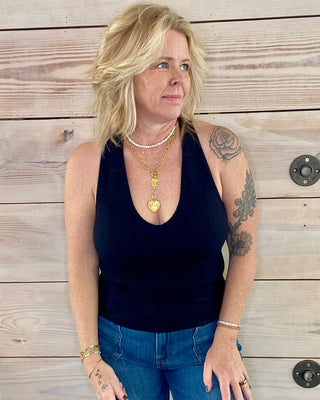 Woman wearing a black tank top and blue jeans standing against a wooden wall.