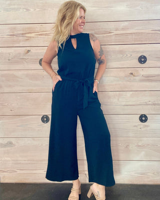 Woman wearing a  black jumpsuit standing against a wooden wall.