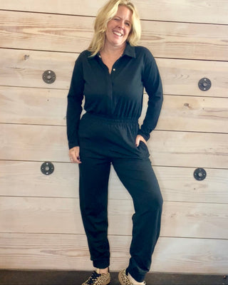 Woman wearing a black jumpsuit standing against a wooden wall.