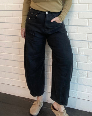 Woman wearing a beige long-sleeve top and black pants against a white brick wall.