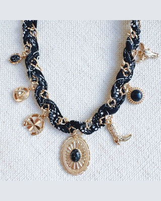 A gothic-style charm necklace featuring a black and white patterned braided cord, gold-tone chain links, and various charms including a large sunburst medallion, crescent moon, and small black gemstones, displayed on a textured white fabric background.