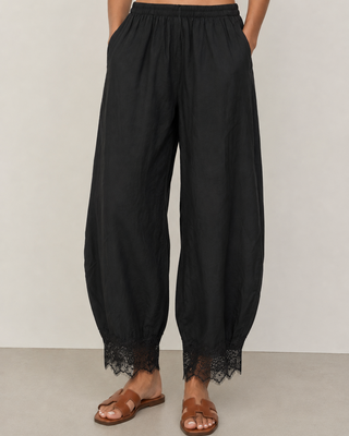 Black wide-leg harem pants with a lace hem, shown on a model wearing brown leather slide sandals against a neutral studio background.