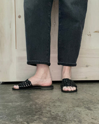 Person wearing black sandals and dark jeans standing on a concrete floor with a wooden door in the background.