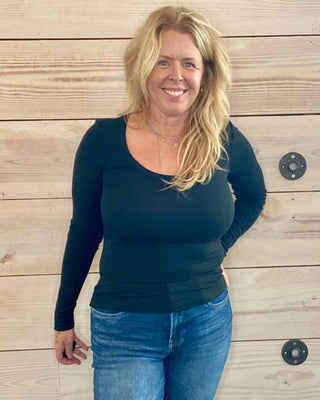 Woman wearing a black long-sleeve top and blue jeans standing against a wooden wall.