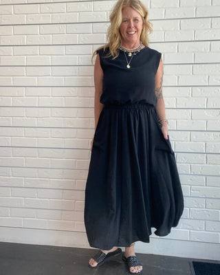A woman wearing a sleeveless black midi dress with a gathered waist and side pockets, standing against a white brick wall.