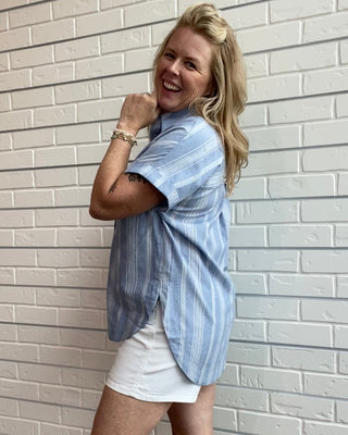 Woman wearing a blue striped shirt and white shorts against a light brick wall.