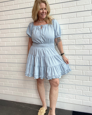 Woman wearing a light blue dress standing against a white brick wall.