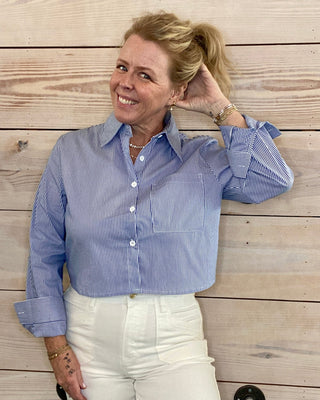 Woman wearing a blue striped shirt and white pants against a wooden wall.