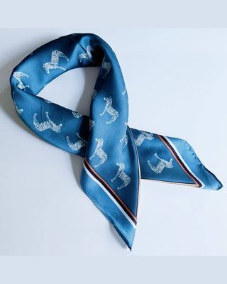 A blue silk scarf with a repeating white zebra print and striped borders, styled in a loop on a light gray background.