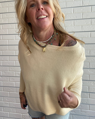 A smiling blonde woman wearing a cream-colored off-the-shoulder waffle-knit sweater and light denim shorts, standing against a white brick wall.