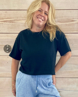 Person wearing a black t-shirt and blue pants standing against a wooden wall.