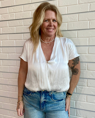 Woman wearing a white blouse and blue jeans standing against a light brick wall.
