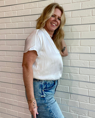 Woman wearing a white top and blue jeans leaning against a light brick wall.