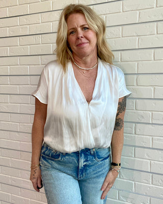 Woman wearing a white blouse and blue jeans standing against a light brick wall.