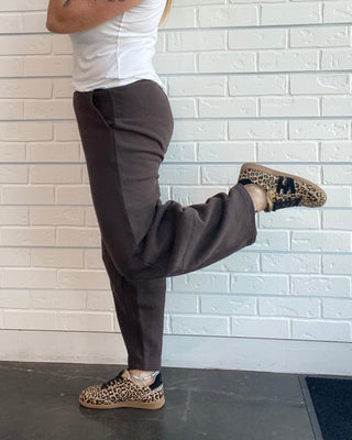 A woman posing in a white tank top, dark brown wide-leg sweatpants, and leopard-print sneakers against a white brick wall.