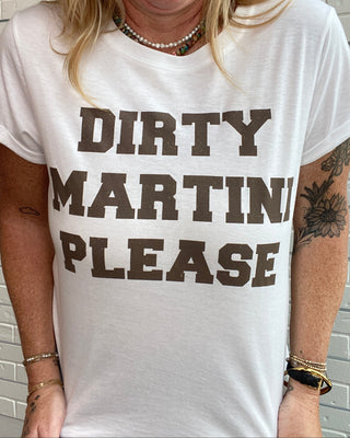 A woman wearing a white graphic t-shirt with the words 'DIRTY MARTINI PLEASE' printed in dark brown block letters, standing against a white brick wall.