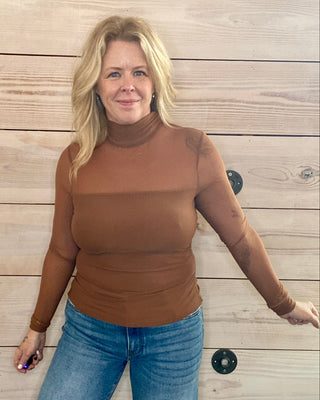 Woman wearing a brown turtleneck and blue jeans standing against a wooden wall.