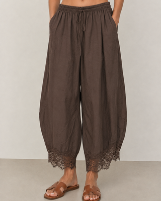 A person wearing loose, drawstring harem-style pants in a dark brown color with lace trim at the hems, paired with brown slide sandals, against a plain light gray studio background.