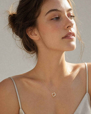 A close-up lifestyle shot of a woman wearing a delicate gold necklace with a small geometric pendant, set against a neutral light gray background.