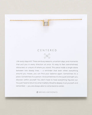 A gold necklace with a square bezel-set clear stone is displayed on a white card that reads 'CENTERED' with a quote by Bryan Anthony. The card is photographed against a light beige background.