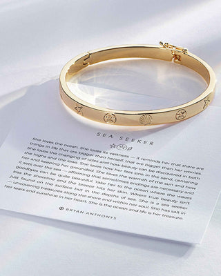 A gold Sea Seeker hinged bangle bracelet with engraved sea creature motifs, displayed on top of its printed paper card featuring the 'Sea Seeker' quote and the brand name Bryan Anthony's.
