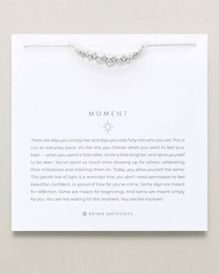 A silver-toned crystal necklace displayed on a white card from Bryan Anthony's, featuring inspirational text under the heading 'MOMENT'.