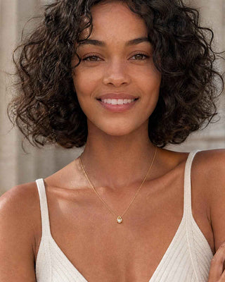 A smiling woman with short curly dark hair wears a delicate gold necklace with a small teardrop pendant, paired with a white ribbed spaghetti-strap top against a soft neutral background.