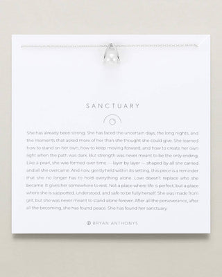 A white rectangular memorial card with the title 'SANCTUARY' and a block of personalized text, suspended from a clear glass bead on a neutral beige background.