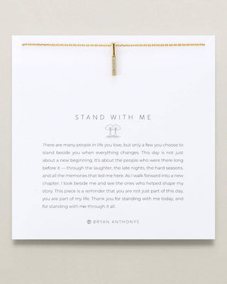 A white square card with the words 'STAND WITH ME' and a short memorial-style message, clipped at the top by a delicate gold necklace. The card is photographed against a light warm neutral background.