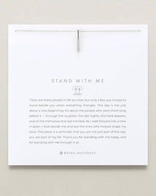 A square white card with a silver hanging ribbon and printed message reading 'STAND WITH ME' by Bryan Anthony's, shown against a light beige background.