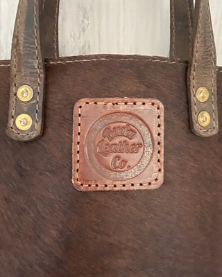 Close-up of a brown leather bag with a central embossed patch reading 'Ruda Leather Co.' and brass-tone rivets on the strap attachments.