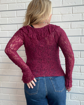 A woman viewed from behind wearing a burgundy lace long-sleeve top and blue jeans, standing against a white painted brick wall.