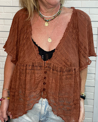 A woman wearing a brown textured short-sleeve button-front cardigan or shrug over a black top and light denim jeans, standing against a white brick wall.