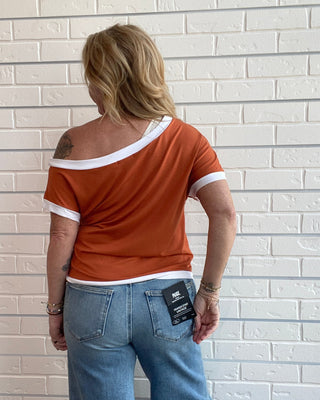 A woman viewed from behind wearing an orange ringer t-shirt with white trim and light blue jeans, standing against a white brick wall.