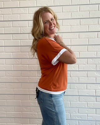 A woman with blonde hair stands against a white brick wall, wearing a rust-orange short-sleeve t-shirt with white contrast trim on the collar and sleeve cuffs, paired with blue jeans.