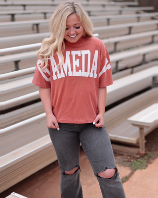 A woman stands in front of outdoor stadium bleachers wearing a terracotta short-sleeve graphic T-shirt with 'CAMEDA' printed in white across the chest, paired with distressed gray jeans.